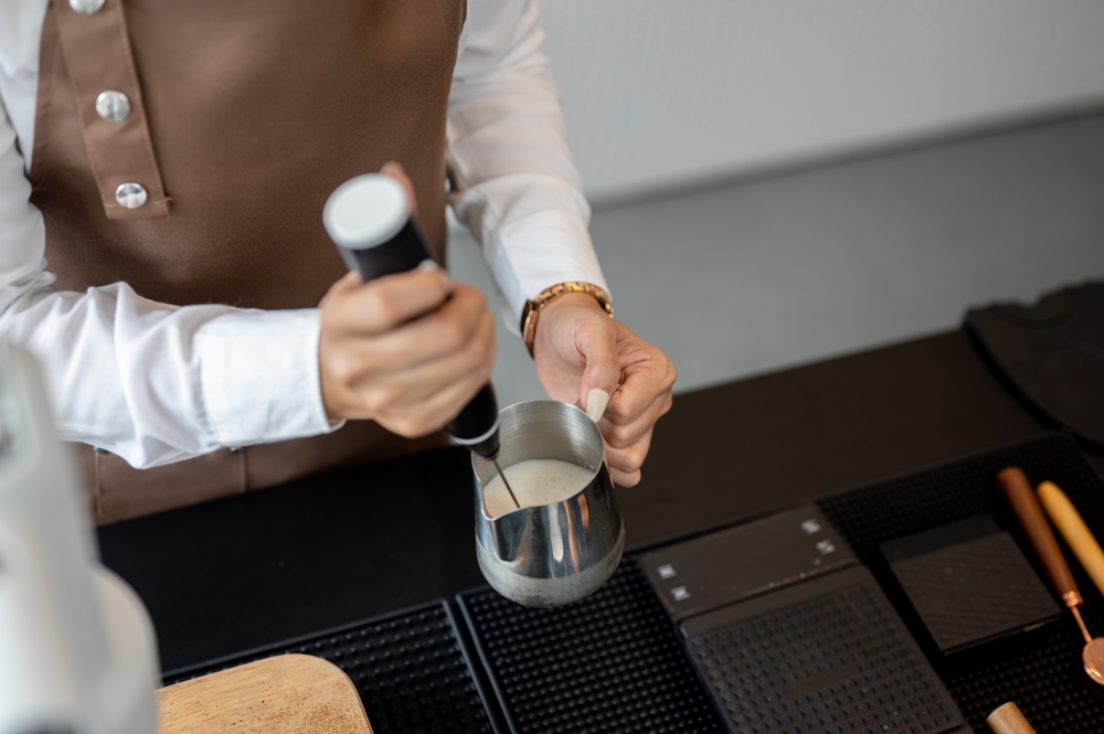 A close-up image of a female barista in an apron using a milk frother blender, making a coffee for customers in a cafe.