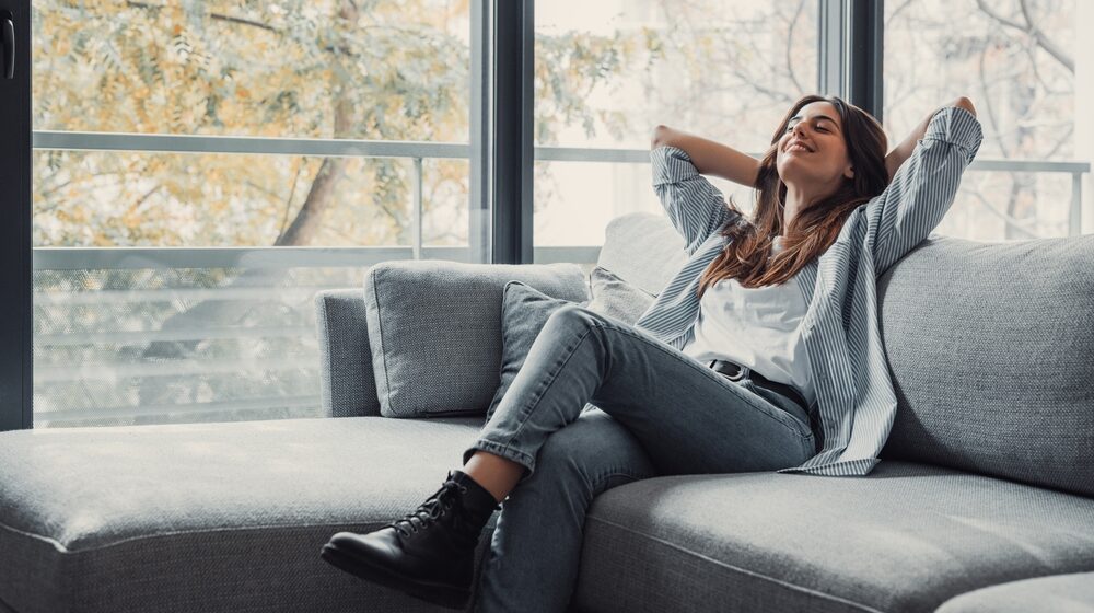 A woman comfortably seated on a couch, with her arms lifted, expressing a feeling of joy or contentment in her space.