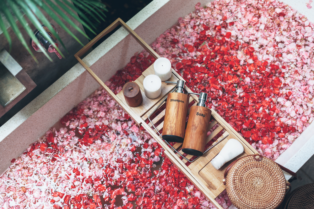 Bathtub with flower petals and beauty products on wooden tray. Organic spa relaxation in luxury Bali outdoor bath.