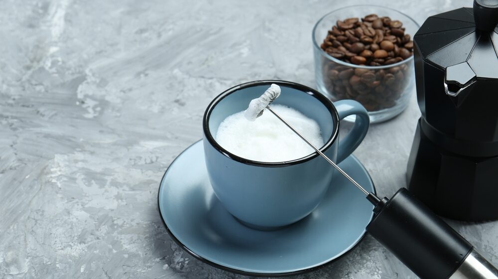 Mini mixer (milk frother), whipped milk in cup and coffee beans on grey textured table, closeup.