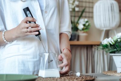 Mixing milk in glass pot by frother closeup. Female making foamy milk with handheld mixer. Housewife woman in home clothes using mixer whips cooking food in kitchen. Cook concept.