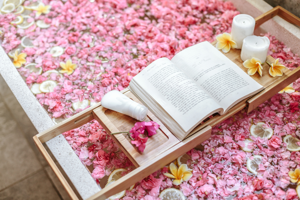 Bath tub with flower petals and lemon slices. Book, candles and beauty product on a tray. Organic spa relaxation in luxury Bali outdoor bathroom.