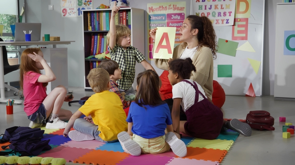 Female teacher hold flashcard with letter a teaching preschool kids alphabet. Diverse pupils of primary school sit on floor with teacher having alphabet lesson