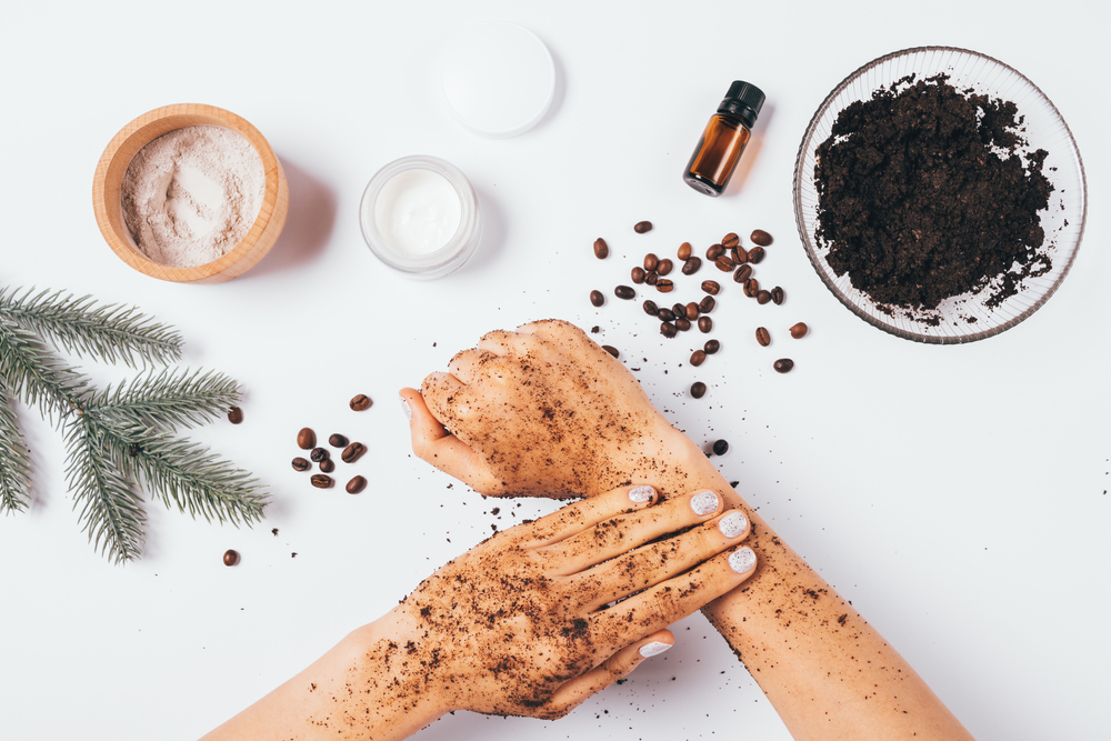 Flat lay female hands making peeling procedure with homemade cosmetic coffee scrub with clay and pine essential oil next to ingredients on white table.