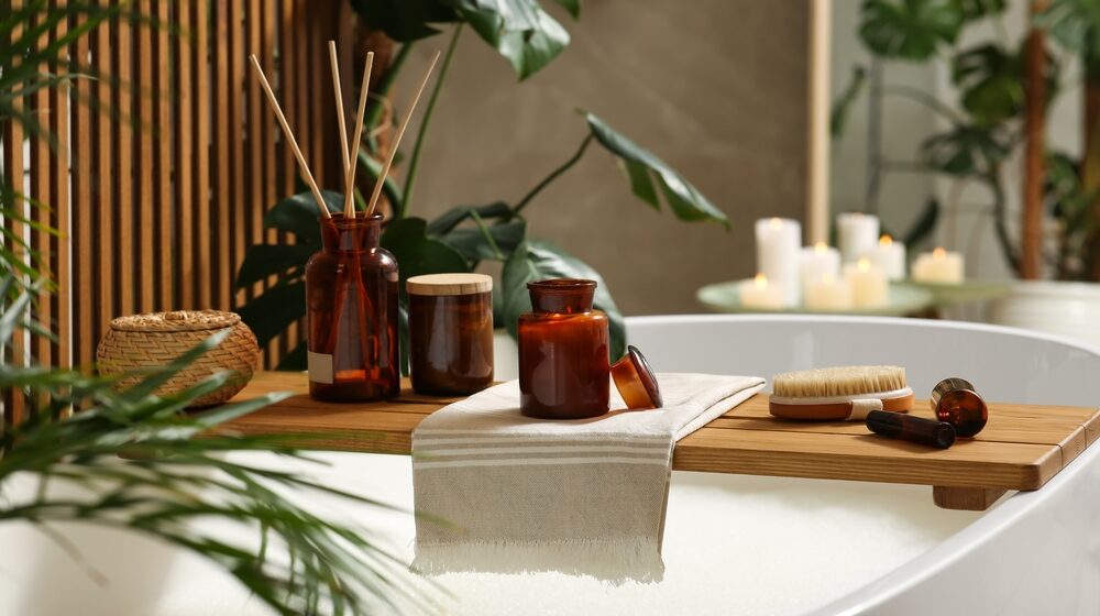 Wooden bath tray with open book candle and body care products on tub indoors. Relaxing atmosphere e1727939801640