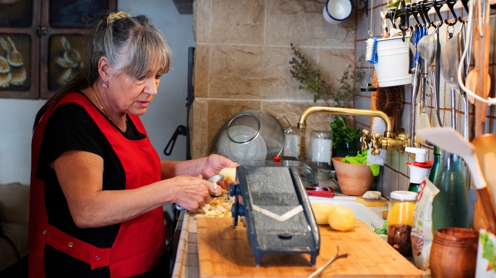 Woman cooking in rustic kitchen, retired person in her sixties with long grey hair, wearing red apron, mandolin for cutting potatoes on top of wood