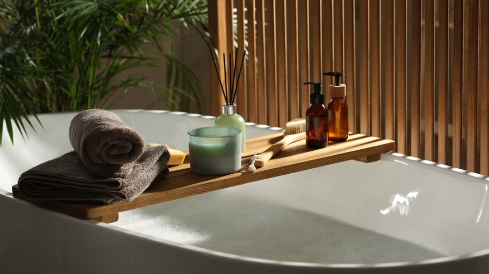 Wooden bath tray with candle, air freshener and bathroom amenities on tub indoors