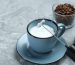 Mini mixer (milk frother), whipped milk in cup and coffee beans on grey textured table, closeup.