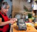 Woman cooking in rustic kitchen, retired person in her sixties with long grey hair, wearing red apron, mandolin for cutting potatoes on top of wood